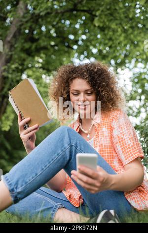 vue en contre-plongée d'une jeune femme souriante avec un ordinateur portable fermé tout en utilisant un smartphone dans un parc vert, image de stock Banque D'Images