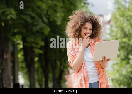 bonne femme indépendante souriant tout en tenant un ordinateur portable et travaillant à distance dans le parc, image de stock Banque D'Images