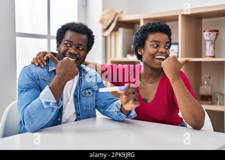 Un jeune couple afro-américain qui tient le résultat du test de grossesse pointe le pouce vers le côté souriant heureux avec la bouche ouverte Banque D'Images
