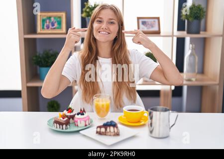 Jeune femme caucasienne mangeant des pâtisseries t pour le petit déjeuner souriant gai montrant et pointant avec les doigts dents et la bouche. concept de santé dentaire. Banque D'Images