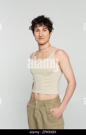 brunette bigender personne dans le réservoir debout avec les mains dans les poches de pantalon beige et sourire à la caméra isolée sur gris, image de stock Banque D'Images