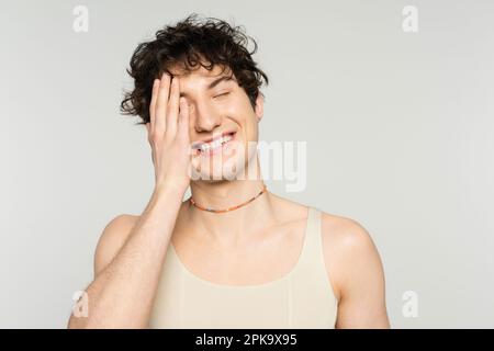 modèle gai non binaire en réservoir et perles couvrant la face avec la main et souriant avec les yeux fermés isolés sur gris, image de stock Banque D'Images