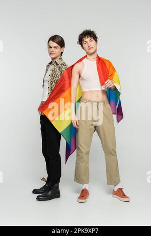 pleine longueur de partenaires pansexués tendance avec drapeau lgbt regardant la caméra tout en se tenant sur fond gris, image de stock Banque D'Images