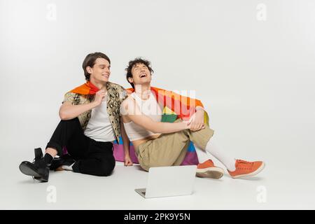 couple pangenre excité avec drapeau arc-en-ciel assis près de l'ordinateur portable sur fond gris, image de stock Banque D'Images