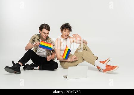heureux couple pansexuel avec drapeau lgbt ayant chat vidéo sur ordinateur portable tout en étant assis sur fond gris, image de stock Banque D'Images