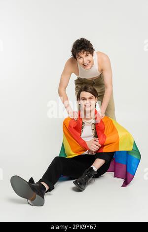 surjoyeuse personne non binaire souriant près de partenaire assis avec un drapeau arc-en-ciel sur fond gris, image de stock Banque D'Images
