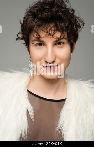 portrait de la personne bigéante positive avec le perçage de nez argenté et la doublure d'oeil arc-en-ciel regardant la caméra isolée sur gris, image de stock Banque D'Images