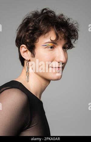 portrait du modèle bigender souriant avec un contour des yeux arc-en-ciel et des accessoires argentés isolés sur une image grise Banque D'Images