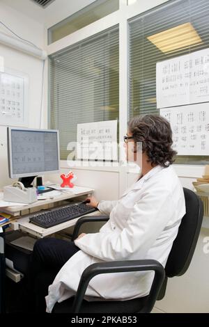 Laboratoire de cytogénétique, entouré de caryotypes, un médecin analyse les résultats d'un échantillon afin de rédiger un rapport. Banque D'Images