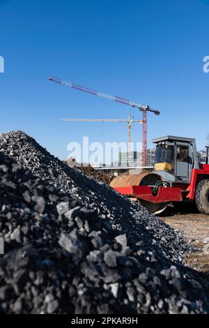 Le rouleau rouge se trouve sur un grand chantier de construction devant des monticules de gravier avec des grues de construction en arrière-plan Banque D'Images