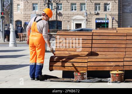 Femme ouvrier en uniforme orange peint le banc en bois sur une rue. Travail de services communautaires, amélioration de la ville au printemps Banque D'Images
