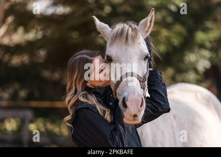 Jeune femme debout à côté des yeux blancs de cheval arabe fermé comme si elle s'embrasse ou sentait, brouillé arbre arrière-plan, gros plan Banque D'Images