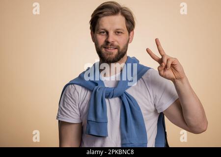 Jeune homme caucasien montrant le signe de la victoire, espérant le succès et la victoire, faisant le geste de la paix, souriant avec une expression aimable et optimiste. Beau gars barbu isolé seul sur fond beige studio Banque D'Images