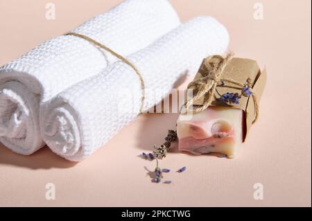 Encore la vie avec des serviettes roulées près de la barre de savon organique naturel et des fleurs de lavande dispersées sur fond rose isolé Banque D'Images