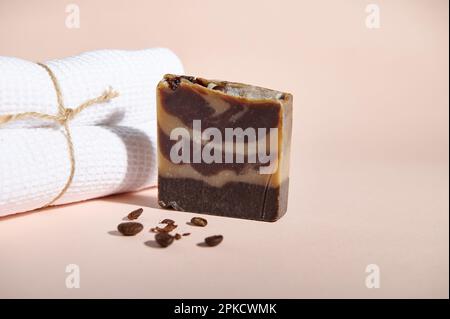 Un bar de savon biologique naturel ou de gommage corporel solide, avec des grains de café moulus à côté d'une serviette de bain blanche sur fond rose Banque D'Images