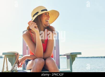 Tenez-vous à votre bonheur. une jeune femme joyeuse portant un chapeau tout en étant assise sur une chaise près d'un lac à l'extérieur au soleil. Banque D'Images