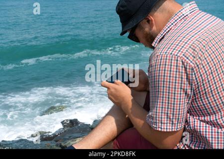 Homme dans une chemise à carreaux assis sur le bord de mer par temps ensoleillé et tenant un téléphone portable dans ses mains Banque D'Images