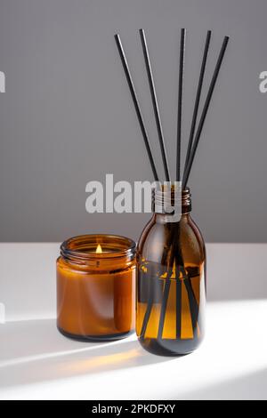 Diffuseur d'arôme avec huile essentielle naturelle dans un flacon de recharge en verre brun, bougie de cire de soja avec feu dans un pot ambre sur fond gris blanc. Banque D'Images