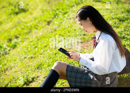 Écolière en uniforme assise sur l'herbe regardant son téléphone Banque D'Images