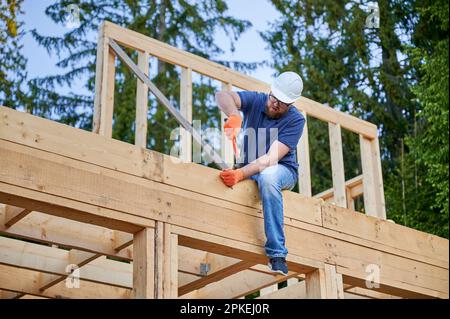 Carpenter construisant une maison en bois de deux étages près des bois. L'homme barbu en lunettes est un clou martelé avec un casque de protection. Le concept de construction écologique moderne. Banque D'Images