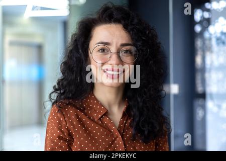 Photo en gros plan d'une femme d'affaires portant des lunettes et des cheveux bouclés souriant et regardant l'appareil photo, portrait d'une femme latino-américaine réussie sur le lieu de travail, fier patron propriétaire d'entreprise. Banque D'Images