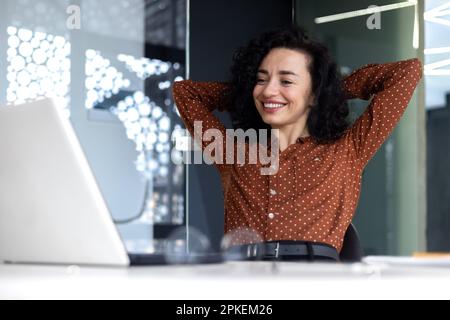 Une femme qui pense bien au travail au bureau, une femme d'affaires qui a les mains derrière la tête a bien terminé son travail, une femme latino-américaine satisfaite des résultats obtenus en regardant l'écran d'un ordinateur portable. Banque D'Images