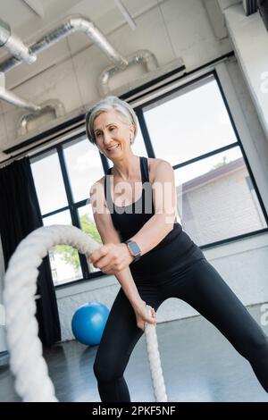 happy senior sportswoman avec cheveux gris s'entraîner avec des cordes de combat dans la salle de gym, image de stock Banque D'Images