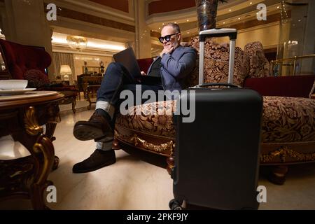 Le voyageur âgé est assis avec un ordinateur portable sur un canapé en velours Banque D'Images