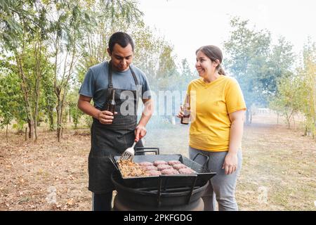 Un jeune couple latin cuisant des hamburgers faits maison sur un barbecue et buvant des bières dans l'arrière-cour. Banque D'Images