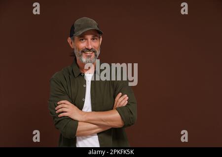 Un homme souriant heureux. Un pêcheur à la casquette de baseball et à la chemise verte se dresse à pattes croisées sur fond marron avec espace pour les copies. Photo de haute qualité Banque D'Images