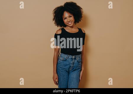 Photo d'intérieur de la jolie femme afro-américaine souriante dans une tenue décontractée regardant l'appareil photo avec un sourire timide et charmant, posant isolée sur le dos beige foncé Banque D'Images