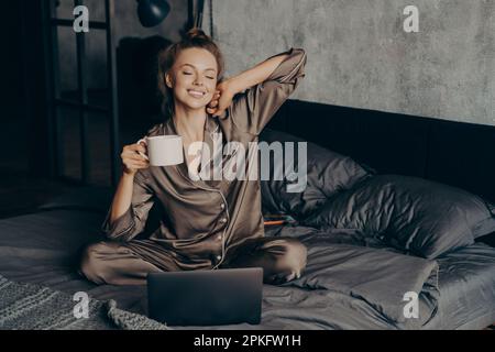 Une femme indépendante décontractée se réveille au lit tout en consultant ses nouveaux messages et e-mails sur son ordinateur portable, vêtue de pyjama. WOM Banque D'Images