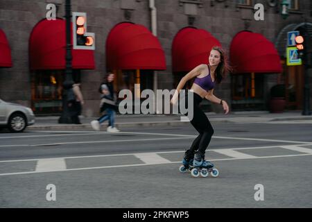 La jeune femme sportive et mince utilise des patins à roulettes comme mode de transport en ville apprécie les passe-temps favoris et stimule l'humeur habillée dans les poses de vêtements actifs sur la route Banque D'Images