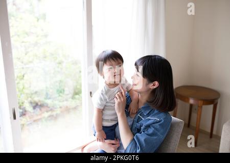 Une mère se détend dans une chaise et un petit garçon s'assoit sur ses genoux dans un salon ensoleillé Banque D'Images