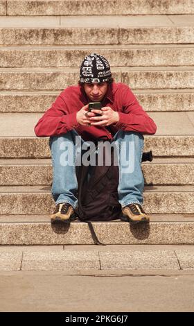 Jeune homme assis sur des marches de pierre à Trafalgar Square, Londres. Royaume-Uni regardant son téléphone portant un chapeau noir et blanc laineux, un pull rouge et un Jean Banque D'Images