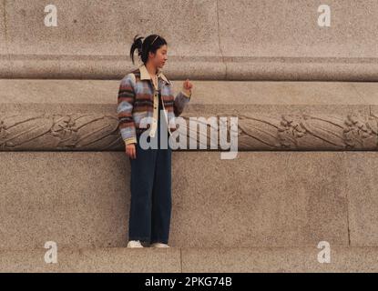 Une jeune femme d'Asie de l'est debout seule sur la plinthe entourant la colonne de Nelson à Trafalgar Square, Londres, Angleterre. ROYAUME-UNI Banque D'Images