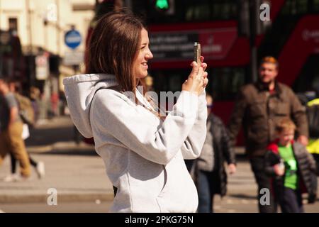 Une jeune femme prenant un selfie inTrafalgar Square, Londres. Royaume-Uni portant un sweat à capuche blanc au soleil Banque D'Images