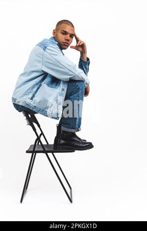 Jeune et beau homme afro-américain dans des vêtements en denim frais assis à l'arrière de la chaise et à l'air sérieux, studio tourné à la verticale. Photo de haute qualité Banque D'Images