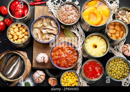 Divers légumes en conserve, viande, poisson et fruits dans des boîtes de conserve. Sur fond sombre. Vue de dessus. Banque D'Images