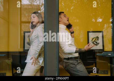 Deux collègues masculins et féminins attrayants ayant un appel téléphonique dans les cabines téléphoniques au bureau Banque D'Images