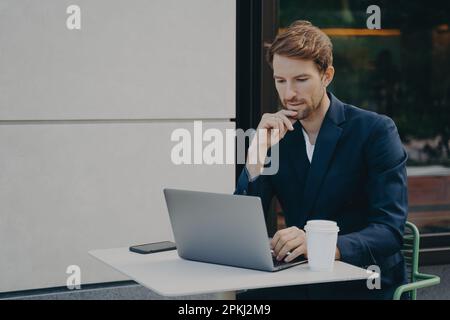 Beau pensif jeune homme freelancer surfant sur le Web sur ordinateur portable tout en étant assis dans un café-terrasse avec la main sur son menton, boire du café et faire en ligne re Banque D'Images