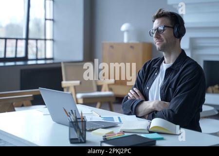 Un employé de banque allemande positif assis sur son lieu de travail dans un bureau à domicile moderne avec un casque sans fil tout en utilisant un ordinateur portable travaillant à distance en ligne, lookin Banque D'Images