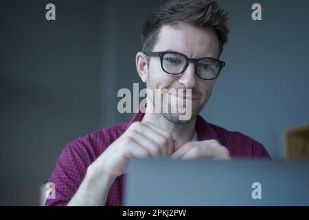 Souriant jeune Allemand indépendant dans les lunettes regardant le rapport de recherche de marketing sur l'écran d'ordinateur portable, l'analyse des données statistiques et le développement de la croissance Banque D'Images