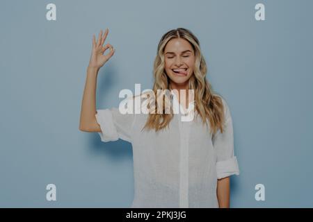 Jeune blonde joyeuse femme caucasienne manifestant un accord et montrant un geste correct avec les yeux fermés, en collant sa langue et en faisant face e drôle Banque D'Images