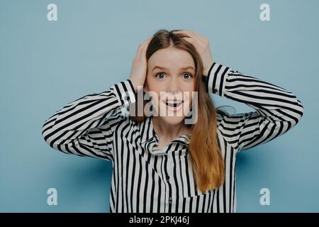 Surprise heureuse adolescente debout en studio, tenant la tête dans les mains et regardant la caméra avec l'expression du visage surprise et la bouche ouverte, femelle b Banque D'Images