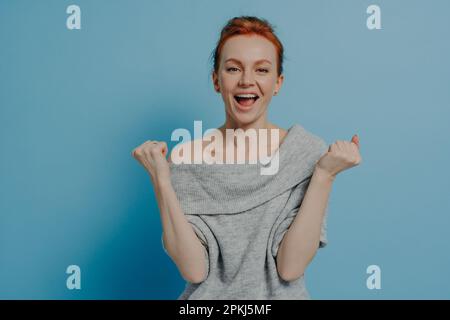 Jeune femme aux cheveux rouges excitée et enjouée vêtue de chandail gris en tricot, qui enragea les poings avec le triomphe et hurle de l'excitation, célébrant le victo Banque D'Images