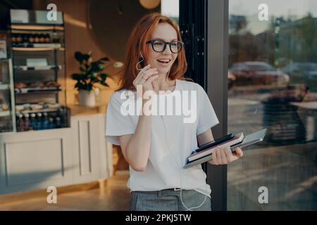 Bonne femme d'affaires aux cheveux rouges avec un ordinateur portable à la main ayant l'appel sur le téléphone mobile, quittant le café, souriant femme dans les lunettes à l'aide d'écouteurs et Banque D'Images