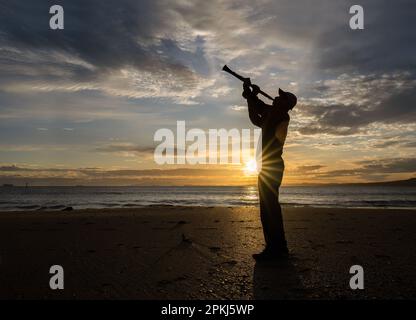 Musicien jouant de la clarinette sur une plage de sable au lever du soleil. Le soleil brille à travers les nuages. Auckland. Banque D'Images