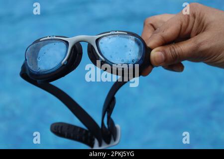 Lunettes de natation pour enfants tenues à la main avec une piscine à l'arrière-plan montrant le concept des activités de vacances d'été Banque D'Images