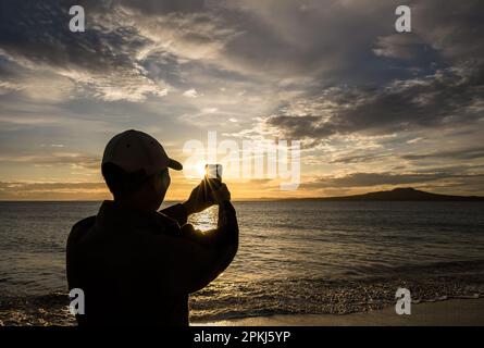 Homme tenant un smartphone et prenant des photos de l'île Rangitoto au lever du soleil. Le soleil brille à travers le bord du téléphone. Auckland. Banque D'Images